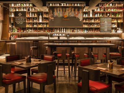 Gemütliche Bar mit dunklem Holz, roten Barhockern und zahlreichen Spirituosenflaschen im Regal.