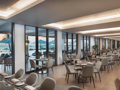 Modernes Restaurant mit großen Fenstern, weißen Tischen, grauen Stühlen und Blick auf das Meer bei Sonnenuntergang.