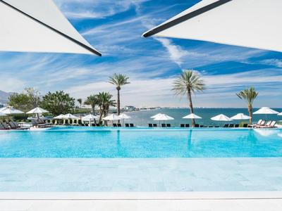 Großes blauer Pool mit weißen Sonnenschirmen, Palmen und Bergen im Hintergrund unter blauem Himmel.