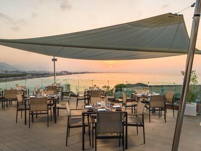 Terrasse mit Tischen und Stühlen unter Sonnensegel, Blick auf Meer und Sonnenuntergang.