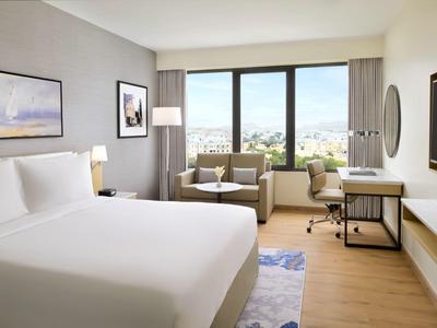 Helles Hotelzimmer mit großem Bett, Sesseln, Schreibtisch und großem Fenster mit Stadtblick.