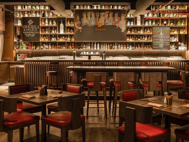 Gemütliche Bar mit dunklem Holz, roten Barhockern und zahlreichen Spirituosenflaschen im Regal.