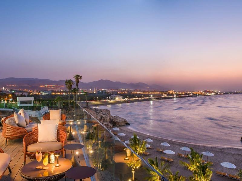 Strandpromenade bei Sonnenuntergang mit Sitzbereichen, Palmen und ruhigem Meer im Hintergrund.