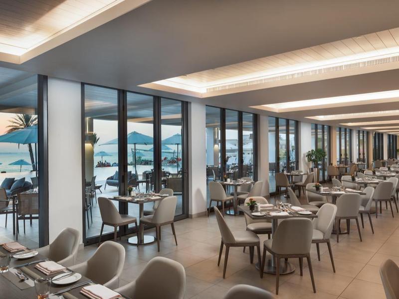 Modernes Restaurant mit großen Fenstern, weißen Tischen, grauen Stühlen und Blick auf das Meer bei Sonnenuntergang.