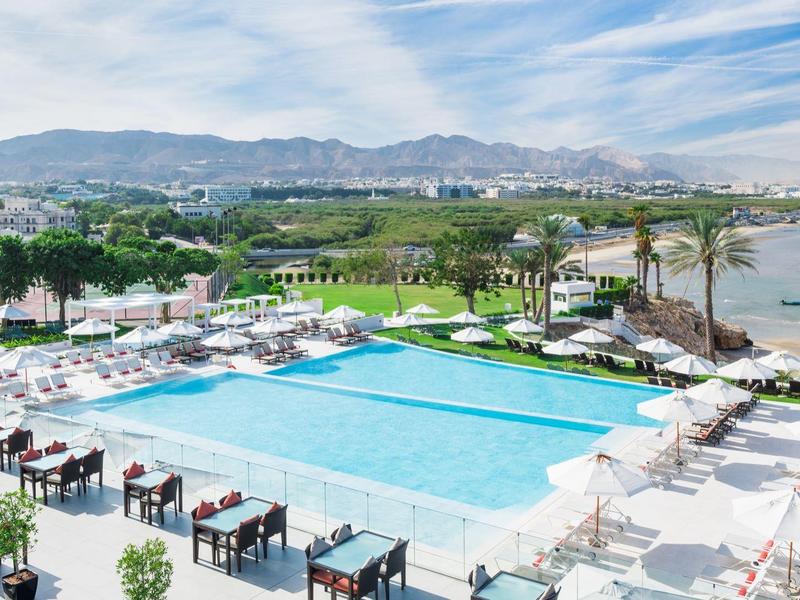 Großer Außenpool mit Liegestühlen und Tischen, Palmen, Blick auf See und Berge im Hintergrund.