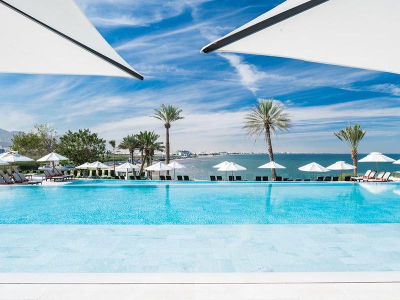 Großes blauer Pool mit weißen Sonnenschirmen, Palmen und Bergen im Hintergrund unter blauem Himmel.