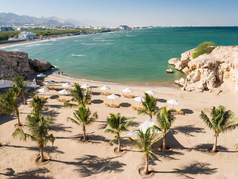 Strand mit Palmen, Sand, Sonnenschirmen und türkisblauem Meer unter klarem Himmel.