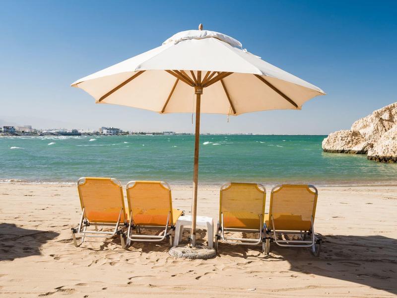 Vier Liegestühle mit weißem Sonnenschirm am Sandstrand vor blauem Meer und klarem Himmel.