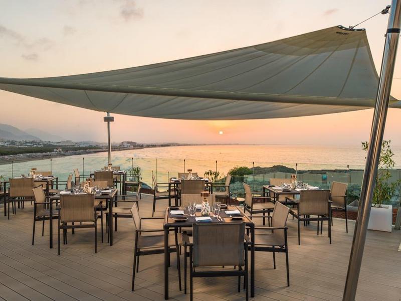 Terrasse mit Tischen und Stühlen unter Sonnensegel, Blick auf Meer und Sonnenuntergang.