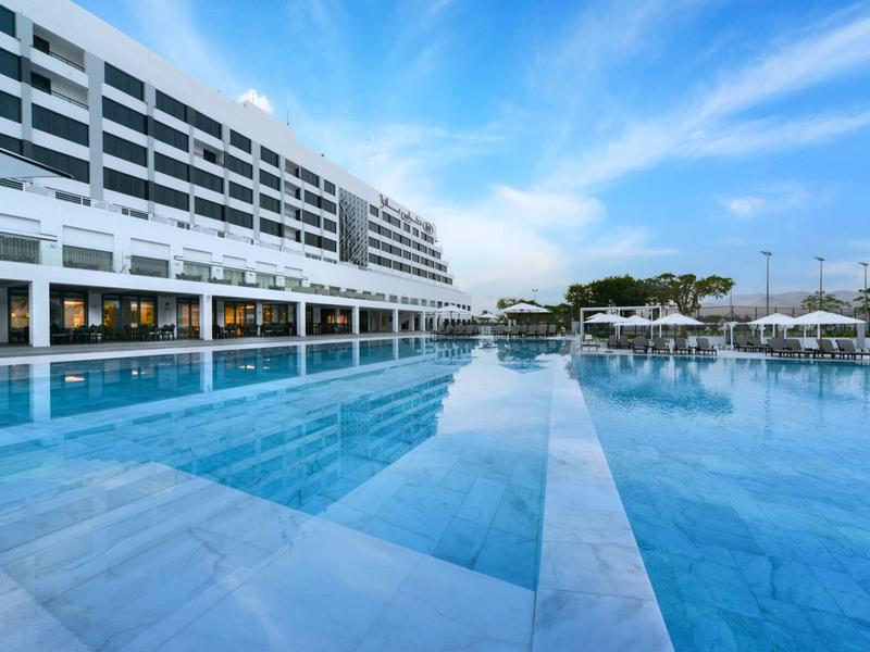 Moderner Hotelbau mit großem, glasklarem Swimmingpool unter blauem Himmel.