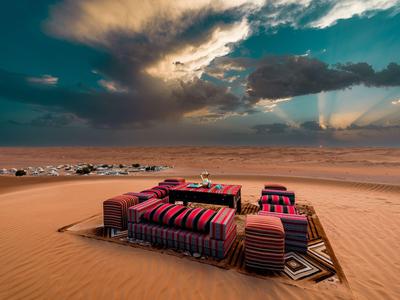 Gemütliche Sitzgruppe mit bunten Kissen auf Sand mit dramatischem Himmel und Dünen im Hintergrund.