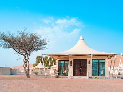 Kleines, weißes Zelt mit spitzem Dach in wüstenartiger Landschaft und blauem Himmel.