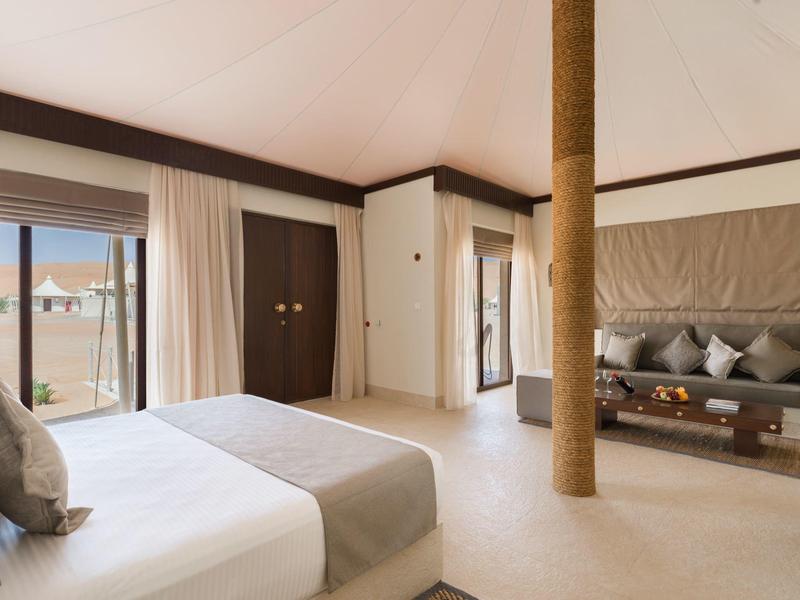 Helles Zimmer mit Doppelbett, großen Fenstern, weißem Vorhang, Säule in der Mitte und Couch mit Kissen.