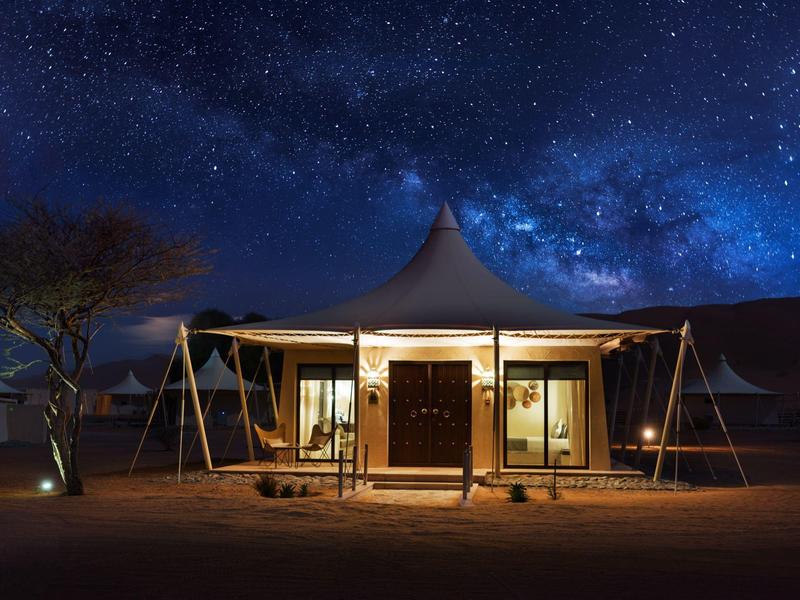 Zelt mit Beleuchtung unter sternklarem Nachthimmel und Milchstraße, umgeben von Wüstenlandschaft.
