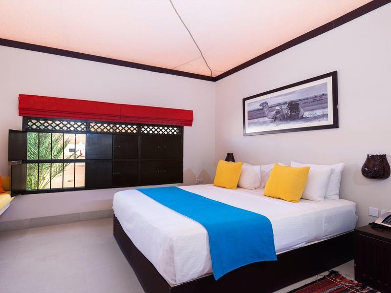 Hell eingerichtetes Schlafzimmer mit großem Bett, blauem Bettläufer, bunten Kissen und Fenster mit rotem Vorhang.