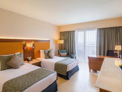 Jasno urządźony pokój hotelowy z dwoma łóżkami pojedynczymi, biurkiem i balkonem.