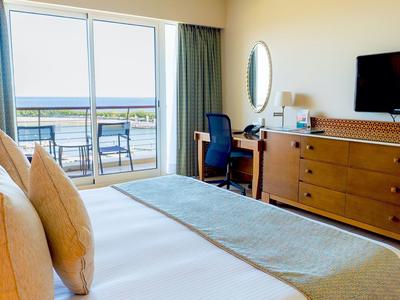 Jasny pokój hotelowy z dużym łóżkiem, biurkiem, telewizorem i balkonem z widokiem na morze.