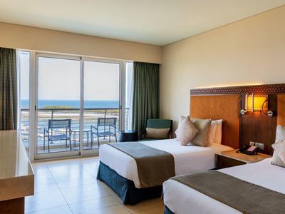 Pokój hotelowy z dwoma pojedynczymi łóżkami, balkonem i widokiem na morze w świetle dziennym.