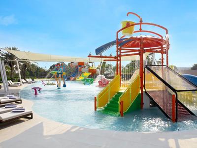 Kolorowy basen dla dzieci z zjeżdżalniami i leżakami pod niebieskim niebem.