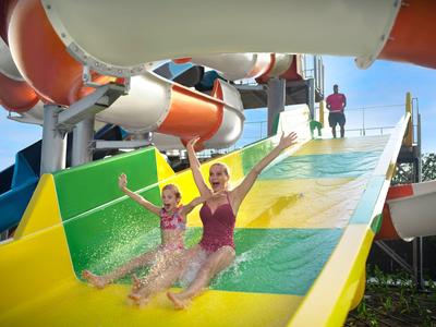 Zwei Kinder rutschen lachend eine bunte Wasserrutsche im Freien hinunter.