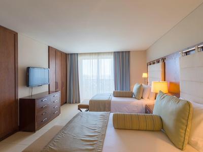 Jasny pokój hotelowy z dwoma łóżkami, sofą, telewizorem i dużą szafą.