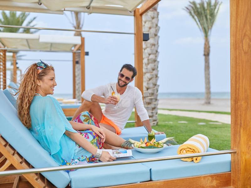 Para relaksuje się na leżakach pod drewnianym baldachimem na plaży z napojami i przekąskami.