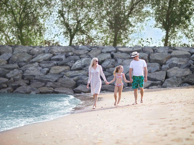 Rodzina z dwojgiem dzieci spacerująca wzdłuż plaży obok kamiennego muru i drzew.