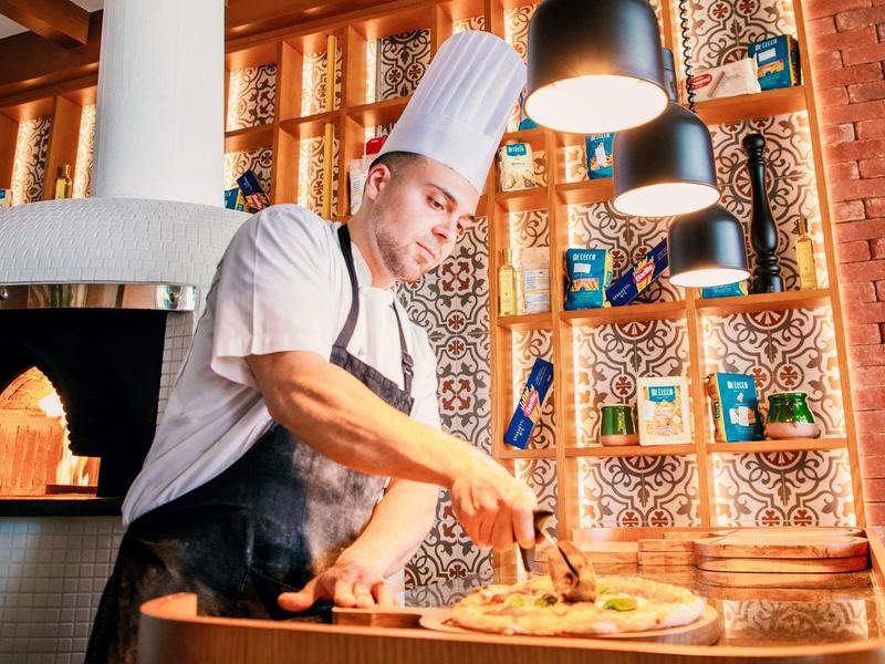 Kucharz w białej czapce przygotowuje pizzę w ciepło oświetlonej restauracji z piecem opalanym drewnem.