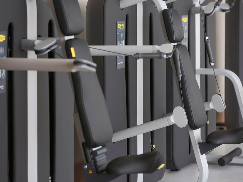 Nowoczesne maszyny fitness w hotelowej siłowni z szarymi poduszkami i metalowymi ramami.