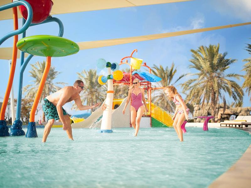 Dzieci bawiące się w płytkim basenie z kolorowymi atrakcjami wodnymi i palmami