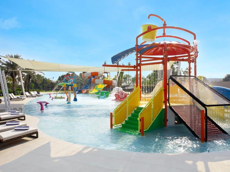 Kolorowy basen dla dzieci z zjeżdżalniami i leżakami pod niebieskim niebem.