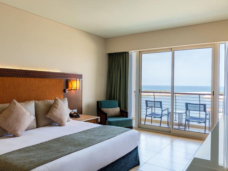 Jasny pokój hotelowy z dużym łóżkiem, miejscem do siedzenia i balkonem z widokiem na morze.