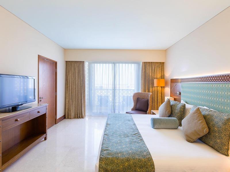 Jasny pokój hotelowy z podwójnym łóżkiem, miejscem do siedzenia, telewizorem i dużym oknem z zasłonami.