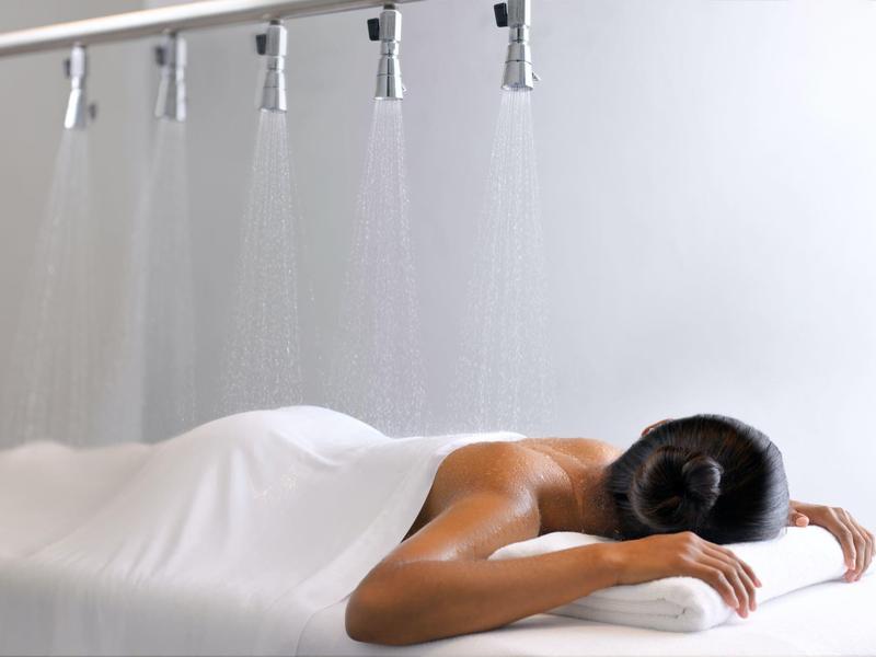 Osoba leží uvolněně na masážním stole pod několika sprchami.