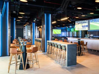 Moderner Barbereich mit hohen Stühlen, blauer Beleuchtung und mehreren Fernsehern an der Wand.