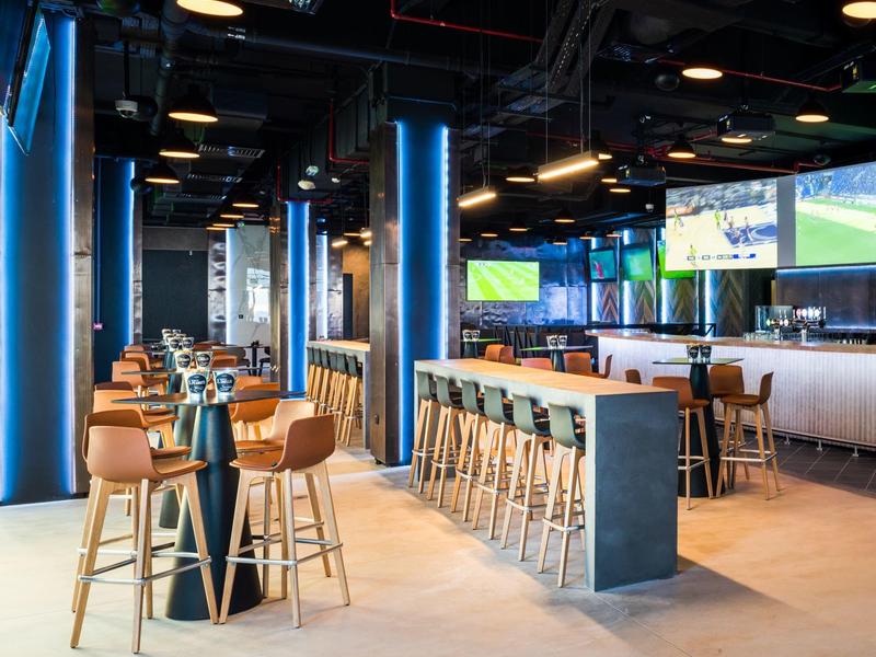 Moderne hotelbar met hoge meubels, blauwe verlichting en meerdere tv-schermen.