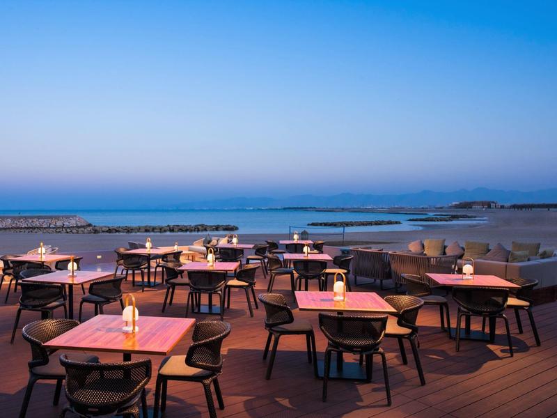 Buitenrestaurant met verlichte tafels op terras bij het strand bij zonsondergang.