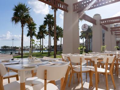 Covered terrace with white tables and wooden chairs, overlooking palm trees and water.