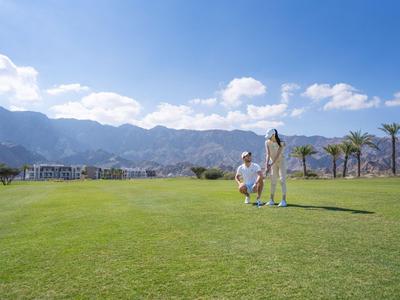Zwei Personen spielen Golf auf einem grünen Feld mit Bergen und Palmen im Hintergrund.