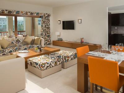 Bright living room with cozy sofas, TV, large windows, and a dining area with orange chairs.