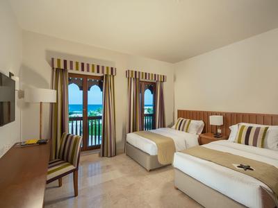 Hotel room with two single beds, desk, chair, and balcony overlooking the sea.