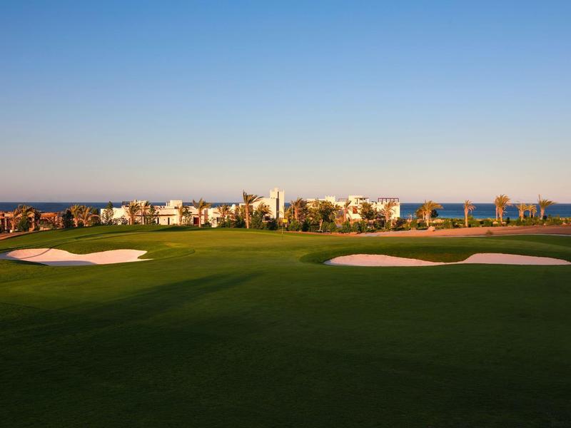 Golf course with manicured green and sand bunkers in front of a village by the sea under clear sky.