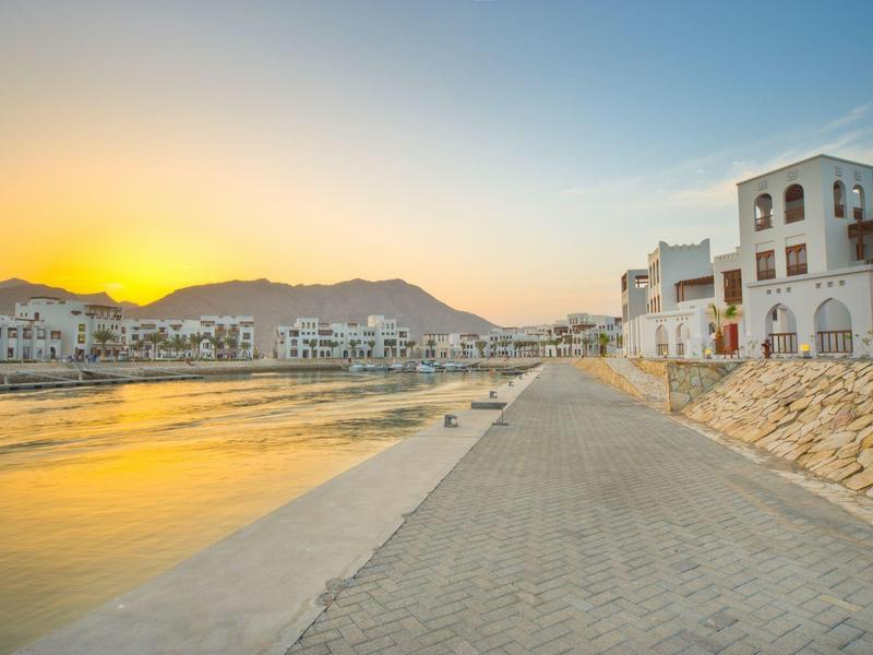 Waterfront promenade with white buildings and mountains in the background at sunset