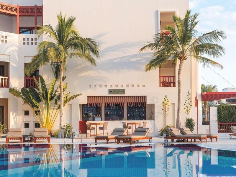 Hotel with pool, sun loungers, and palm trees under clear sky