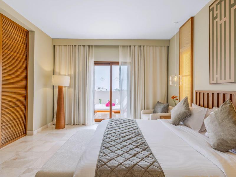 Bright hotel room with large bed, gray bed runner, and balcony access with sea view.