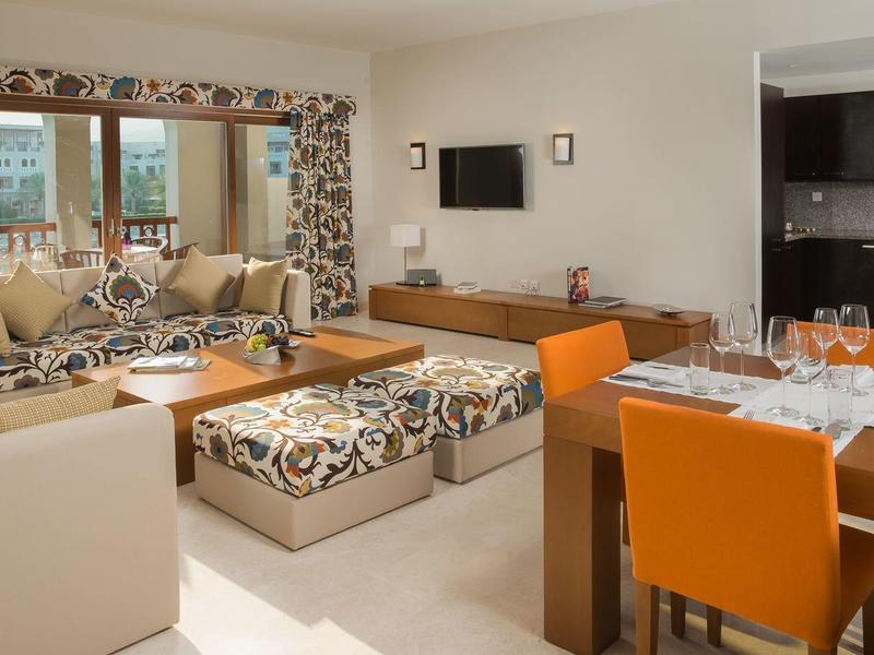 Bright living room with cozy sofas, TV, large windows, and a dining area with orange chairs.