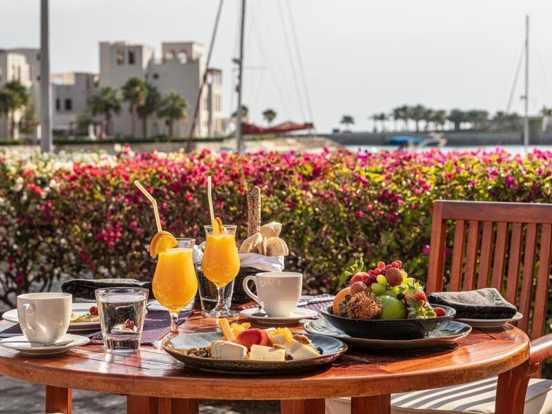 Wooden table on terrace with breakfast, orange juice and view of sea and buildings.