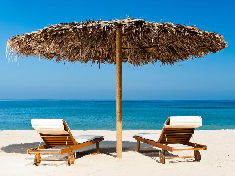 Two wooden lounge chairs with cushions under a straw umbrella on a white sandy beach overlooking the blue sea.