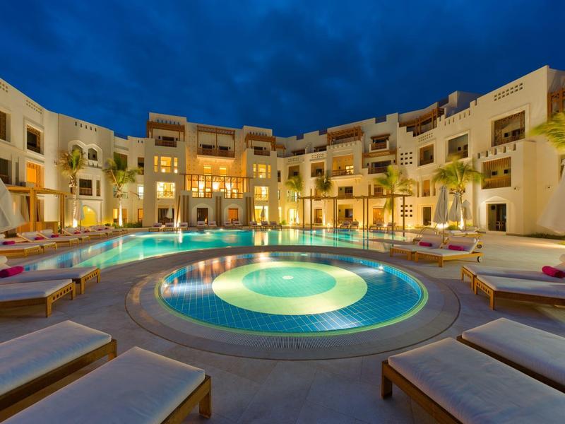 Illuminated hotel pool with loungers and multi-story building in the background at night.