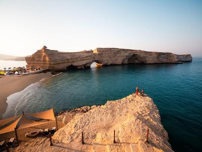 Skalnatý ostrov s přírodním obloukem v moři a skalnaté pobřeží pod jasnou oblohou.